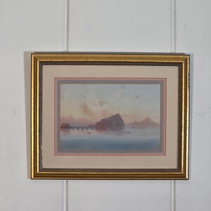 May include: Framed watercolor painting of a coastal landscape. The artwork features a rocky island, a pier, and small boats on the water. The sky has soft pink and blue hues, and the frame is gold.
