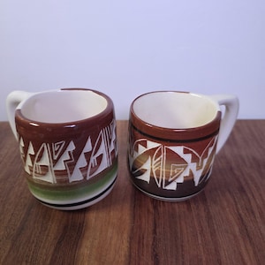 May include: Two ceramic mugs featuring a white interior and a brown exterior with a carved geometric pattern. The left mug has a green band near the base, while the right mug features a dark brown base. Both mugs have a white handle and a rounded shape.