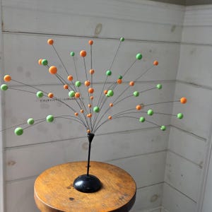 May include: A decorative metal sculpture featuring a black base and stem, with thin wires extending outwards. The wires hold various sized orange and green spheres, creating a burst-like design. The sculpture is placed on a small, round wooden stool.
