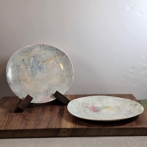 May include: Two round ceramic plates featuring a light blue and white grid pattern with abstract pastel accents in pink, yellow, and blue. One plate is displayed upright on a dark wooden stand, while the other rests flat on a dark wooden surface.
