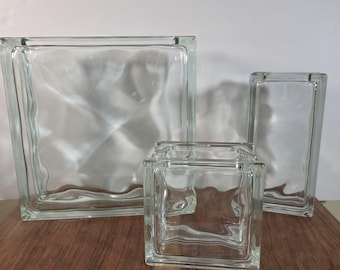 May include: Three clear glass blocks of varying sizes arranged together. The largest is a square, the medium is a smaller square, and the smallest is a tall rectangle. Each block features a wavy, textured surface that distorts light and visibility.