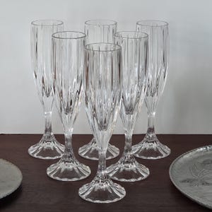 May include: Six clear champagne flutes with vertical ribbed detailing. The glasses are arranged on a dark wooden surface, with a decorative metal plate visible on the left and right sides. The flutes have a classic, elegant design.
