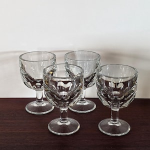 May include: Four clear, stemmed cordial glasses with a decorative, textured pattern on the bowl. The glasses are arranged on a dark wooden surface, with a plain white background.