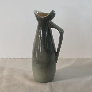 May include: A tall, green ceramic pitcher with a unique handle and spout design. The pitcher has a smooth, glossy finish and a gradient color from dark green to light green. The handle is angular, and the spout has a curved, open design.