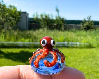 Può includere: Una statuetta di polpo in vetro fatta a mano. Il polpo rosso ha grandi occhi bianchi e poggia su una base di vetro blu con motivi a vortice. La statuetta è tenuta da un dito contro uno sfondo verde sfocato.