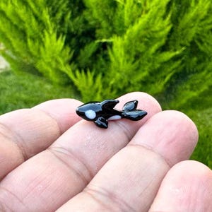 Könnte beinhalten: Eine kleine, handgefertigte Orca-Figur aus Glas. Der Orca ist schwarz mit weißen Markierungen und einem weißen Bauch. Die Figur wird in einer Hand gehalten, mit einem grünen, belaubten Hintergrund.