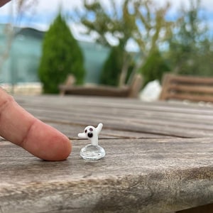 Puede incluir: Una pequeña figura de fantasma de vidrio blanco con ojos negros y una expresión de sorpresa. El fantasma está de pie sobre una base de vidrio transparente.