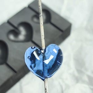 Op de afbeelding: Een blauw hartvormig snoepje aan een stokje. Het snoepje is transparant en heeft een glanzende afwerking. Op de achtergrond is een donkergrijze mal met hartvormige holtes zichtbaar.