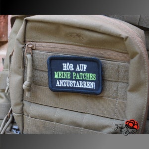 Könnte beinhalten: Ein rechteckiger schwarzer Aufnäher mit weißem und grünem Text, der "HÖR AUF MEINE PATCHES ANZUSTARREN!" lautet. Der Aufnäher ist an einem beigefarbenen taktischen Rucksack mit mehreren Riemen und einem Reißverschluss befestigt.