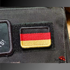 Könnte beinhalten: Nahaufnahme eines rechteckigen Aufnähers mit der deutschen Flagge, mit schwarzen, roten und goldenen horizontalen Streifen. Der Aufnäher ist mit einem goldenen Rand bestickt und auf einem schwarzen Stoff befestigt. Das Bild zeigt die Details des Aufnähers.
