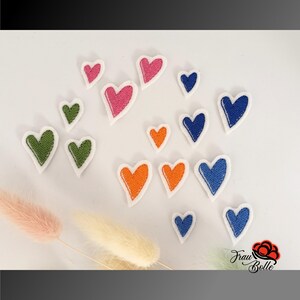 Könnte beinhalten: Eine Sammlung von gestickten Herz-Patches in verschiedenen Farben, darunter Rosa, Orange, Grün und Blau, mit weißen Rändern. Die Patches sind auf einer weißen Oberfläche verstreut, mit getrockneten Blumen im Vordergrund. Die Patches sind in verschiedenen Größen.