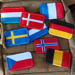 Könnte beinhalten: Eine Sammlung rechteckiger bestickter Aufnäher mit verschiedenen Nationalflaggen, darunter Frankreich, Dänemark, Schweden, Deutschland, Norwegen, Belgien und die Tschechische Republik, angeordnet auf einem beigen Stoff mit Gurtbändern.