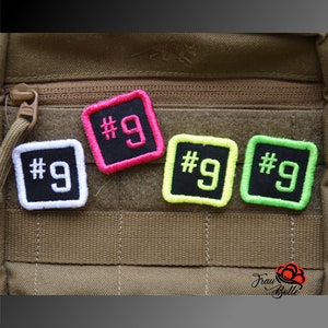 Könnte beinhalten: Vier schwarze quadratische Patches mit weißen, rosa, gelben und grünen Rändern. Jeder Patch hat die Nummer "#9" in weiß auf schwarzem Hintergrund.