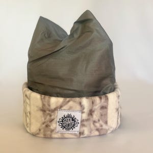 May include: A gray beanie hat with a patterned, cream-colored brim. The brim features a repeating pattern of brown and gray shapes. A small white tag with black text and a sunburst design is sewn onto the brim.