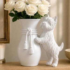 May include: A white ceramic vase featuring a sculpted West Highland White Terrier dog standing on its hind legs against the side. The vase is adorned with a white rope detail tied in a knot with two tassels and contains a bouquet of white roses.