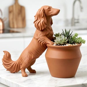 May include: A terracotta-colored dachshund dog figurine leans against a matching terracotta pot filled with succulents and small pebbles. The dog has detailed fur and is posed as if looking at the plants. The pot is round with a slightly flared rim.