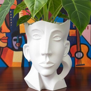 May include: A white ceramic planter shaped like a human head with a textured matte finish. The vessel features stylized facial features including eyes, a nose, and lips, with an open top containing a green leafy plant with large, pointed leaves.
