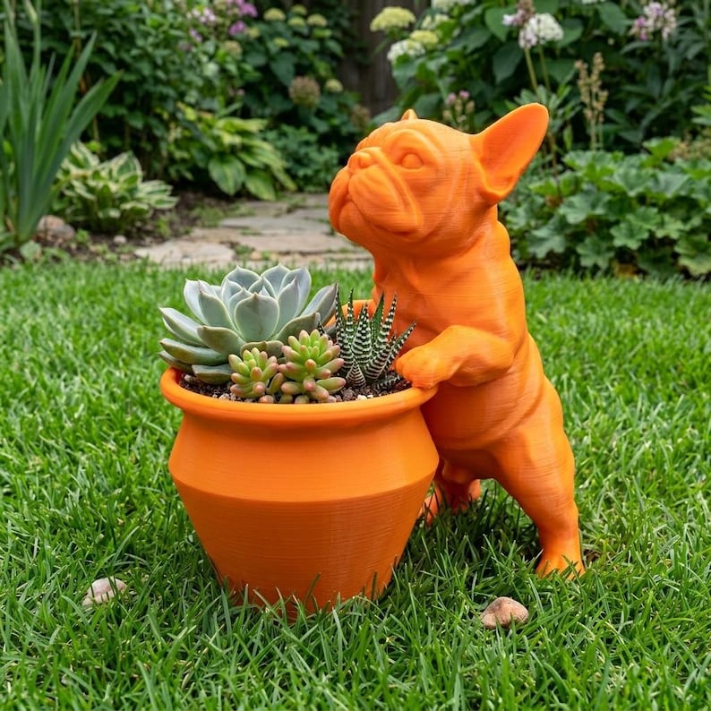 May include: A bright orange planter shaped like a French Bulldog standing on its hind legs with its front paws resting on the rim. The planter contains a variety of small green succulents, including a large rosette-shaped plant and a spiky plant.