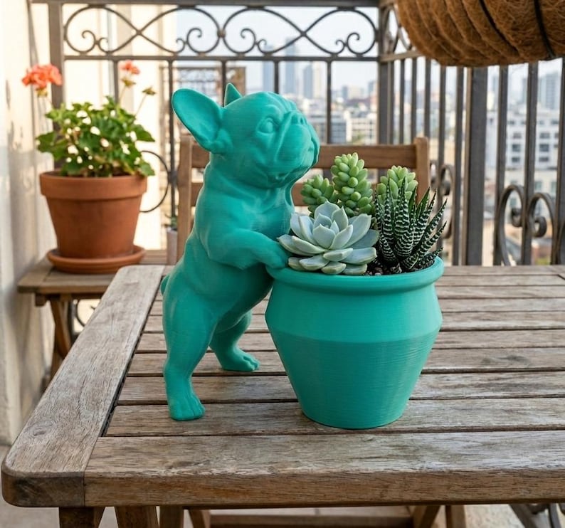 May include: A teal-colored French bulldog figurine standing beside a matching teal planter pot. The planter contains a variety of small green succulents, including a rosette-shaped plant and a spiky, striped succulent with pointed leaves.