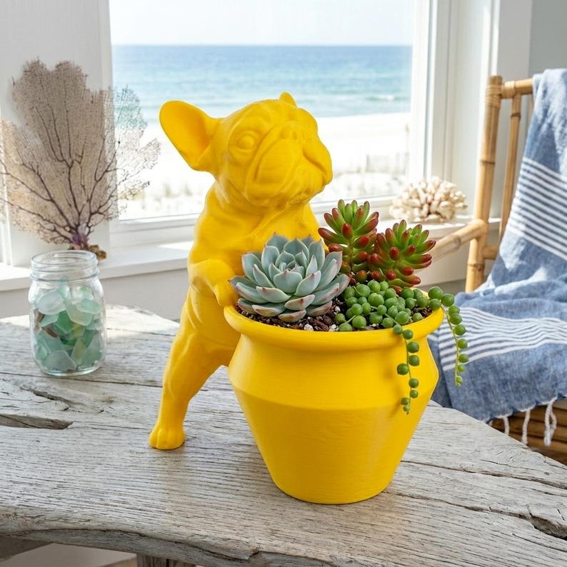 May include: A bright yellow French bulldog figurine standing on its hind legs, holding a matching yellow planter pot filled with a variety of green and reddish succulents, including a large rosette-shaped succulent and trailing string of pearls.