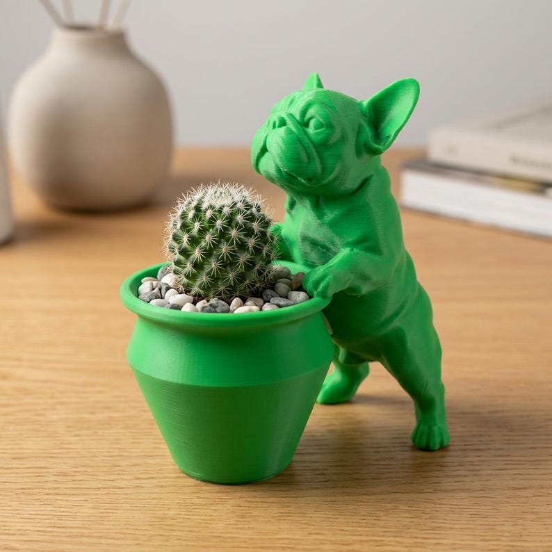 May include: A bright green, 3D-printed French bulldog figurine standing on its hind legs, with its front paws resting on the rim of a matching green planter. The planter contains a small, round, spiky green cactus nestled in gray pebbles.