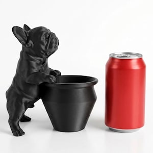 May include: A matte black French bulldog figurine standing on its hind legs with its front paws resting on the rim of a matching black cylindrical planter. A standard red aluminum soda can is positioned next to the planter for scale.