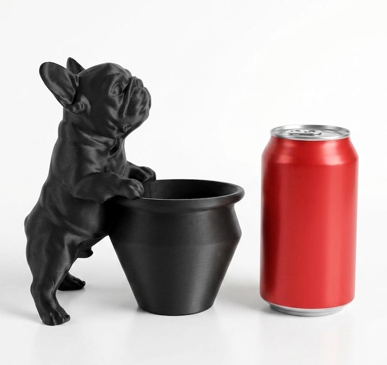 May include: A matte black French bulldog figurine standing on its hind legs with its front paws resting on the rim of a matching black cylindrical planter. A standard red aluminum soda can is positioned next to the planter for scale.