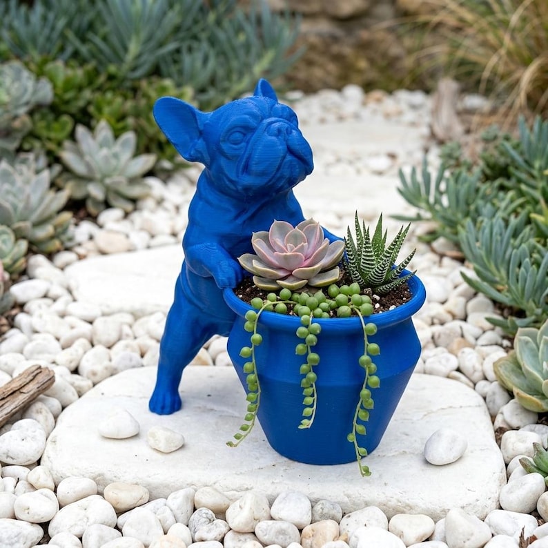 May include: A bright blue French bulldog figurine standing on its hind legs, with its front paws resting on the rim of a matching blue planter. The planter contains a variety of small succulents, including a string of pearls plant.