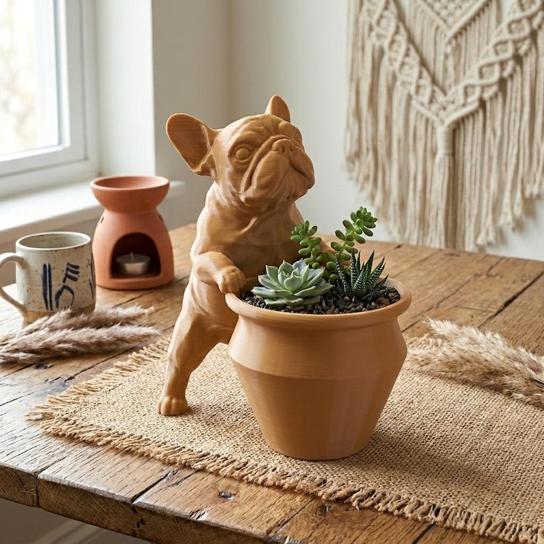 May include: A light brown ceramic planter shaped like a French Bulldog standing on its hind legs, with its front paws resting on the rim of a bowl-shaped pot. The planter contains a small green succulent plant and dark soil, creating a decorative piece.