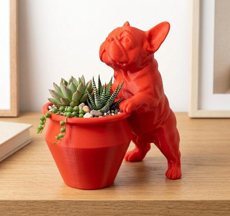 May include: A bright orange planter shaped like a French bulldog standing on its hind legs, with its front paws resting on the rim. The planter contains a variety of small green succulents, including a rosette-shaped plant and a trailing vine.
