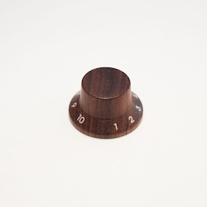 Può includere: Manopola in legno marrone scuro con numeri bianchi da 1 a 10. La manopola ha una parte superiore cilindrica e una base svasata. I numeri sono uniformemente distanziati attorno alla base. La manopola è probabilmente per uno strumento musicale o un dispositivo elettronico.
