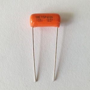 May include: Orange capacitor with two silver leads. The capacitor has the text "SBE 715P600V 103J 1227" printed on it. The component is likely used in electronics.