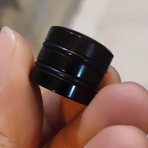 May include: A glossy black bottle stopper with a cylindrical design. The stopper has a flat top and bottom with two raised, circular bands around the middle. The object is held in a person's hand.
