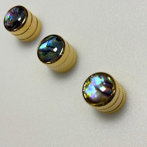 以下が含まれることがあります： テクスチャードデザインの金色のノブが3つ。各ノブは、青、緑、紫の色合いを示す、きらびやかで虹色のあわびの貝殻の象嵌が特徴です。ノブは白い表面に斜めに配置されています。