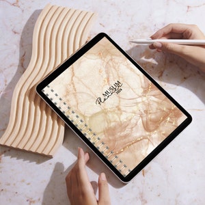Peut inclure: Une tablette numérique avec un design en marbre beige et or. La tablette est ouverte sur un planificateur musulman 2024. La tablette est sur une surface en bois avec un stylet blanc à côté.