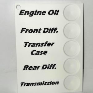 May include: White plastic card with black text and five white circles. The text reads: "Engine Oil", "Front Diff.", "Transfer Case", "Rear Diff.", and "Transmission".