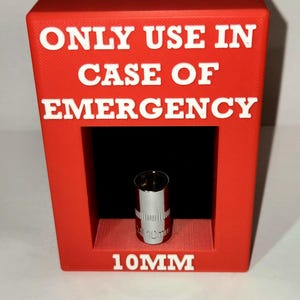 以下が含まれることがあります： 「ONLY USE IN CASE OF EMERGENCY」と「10MM」の文字が書かれた赤いプラスチック製の箱。箱の中に銀色のソケットレンチが入っています。