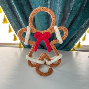 May include: A handmade, crocheted gingerbread man ornament. The ornament features a circular head, arms, and legs, with a red bow tie. The gingerbread man is primarily brown with white and red accents. The ornament is displayed against a teal curtain.