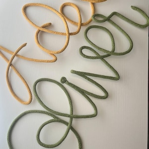 Puede incluir: Un letrero decorativo de pared hecho de cordón tejido, que presenta la palabra 'Hello' en un tono amarillo mostaza y la palabra 'Silver' en un tono verde salvia, ambas escritas en una letra cursiva fluida y continua con bucles.