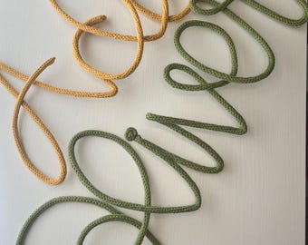 May include: A decorative wall sign crafted from knitted cord, featuring the word 'Hello' in a mustard yellow hue and the word 'Silver' in a sage green shade, both written in a continuous, flowing cursive script with rounded loops.