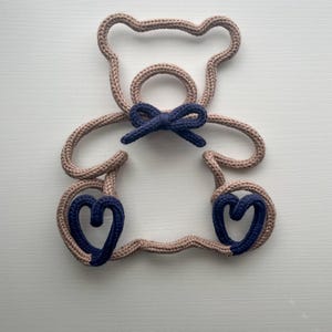 May include: A beige-colored, rope-like teddy bear outline with a navy blue bow and heart-shaped feet. The bear is made of a thick, textured material and is set against a white background. The design is simple and modern, suitable for nursery decor.