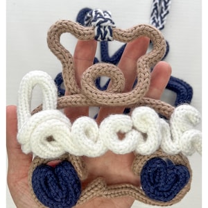 May include: A handmade, crocheted car-shaped ornament in beige, white, and navy blue. The word "boose" is spelled out in white yarn across the front. The wheels are navy blue, and the top is beige. A navy and white rope is attached.