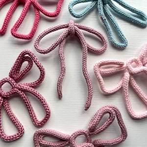 Peut inclure: Gros plan de plusieurs nœuds tricotés dans des tons de rose et de bleu. Les nœuds sont faits d'un fil épais et texturé et sont disposés sur une surface blanche. Les nœuds varient en taille et en forme, certains ayant de longues queues flottantes.