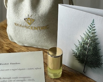 May include: A small bottle of essential oil with a gold cap and a white label with the text "Wooded. Timeless. clean with a rugged elegance - nature, refined." The bottle is sitting on a wooden surface next to a white canvas bag with the text "ECO-SCENTRIC" in gold lettering. A card with a green tree illustration is in the background.