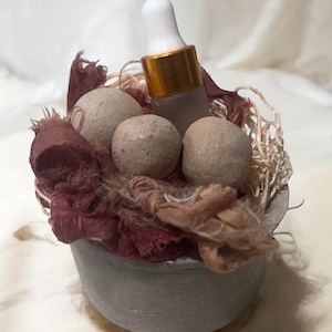 May include: A small concrete planter with a brown fabric lining and four beige stones. A glass bottle with a gold cap and a dropper is nestled in the fabric.