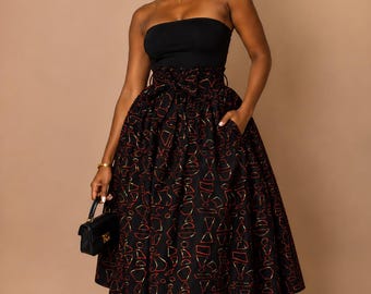 May include: A black strapless top paired with a high-waisted midi skirt featuring a black base and a geometric pattern in red and gold. The outfit includes a matching headwrap, a small black handbag, and black strappy high-heeled sandals.