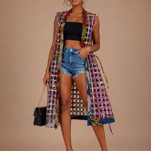 May include: A sleeveless, knee-length duster vest featuring a grid-like cutout pattern with multicolored, patterned fabric strips in shades of blue, red, yellow, and black. The garment is worn over a black tube top and blue denim shorts.