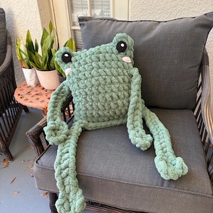 May include: A large, handmade, green crochet frog pillow with black eyes and pink cheeks. The frog is sitting on a gray cushion chair. The frog has long, dangling arms and legs.