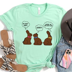 May include: A mint green t-shirt with a graphic of three chocolate bunnies with speech bubbles that read "My butt hurts," "What??" and "Where did you guys go?" The shirt is paired with sandals, jeans, a hat, and a phone.