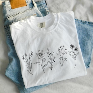 May include: White t-shirt with a black line drawing of a row of flowers on the front. The t-shirt is folded and laid on top of a pair of blue jeans.